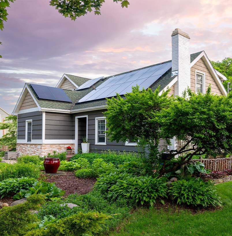 A suburban bungalow features solar panels on the roof and a lush green backyard with various trees and shrubs.