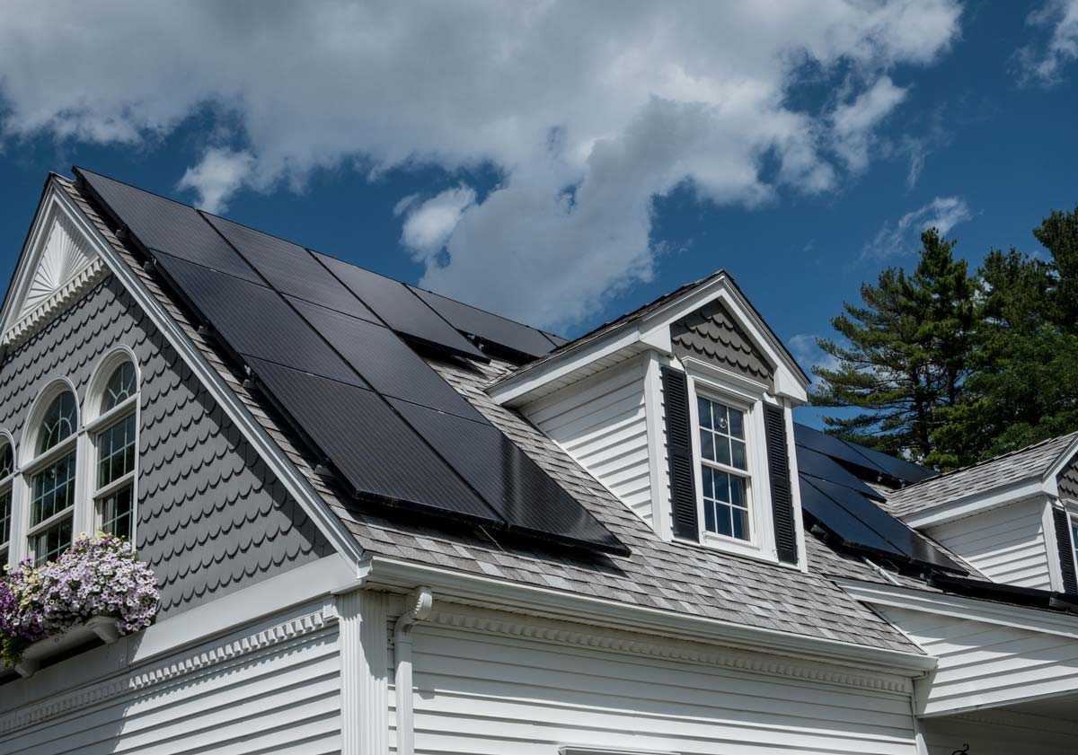 A pristine white home features a dark metal roof with an array of solar panels on top of the roof. The sun sets in the background, casting a glow on the edge of the home and surrounding landscaping.